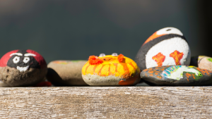 A photo of several painted rocks resting on a wooden fence. Of the ones with their paint visible, one is a red ladybug, one looks like a yellow fish, one has a unicorn painted on it, and another is a puffin.