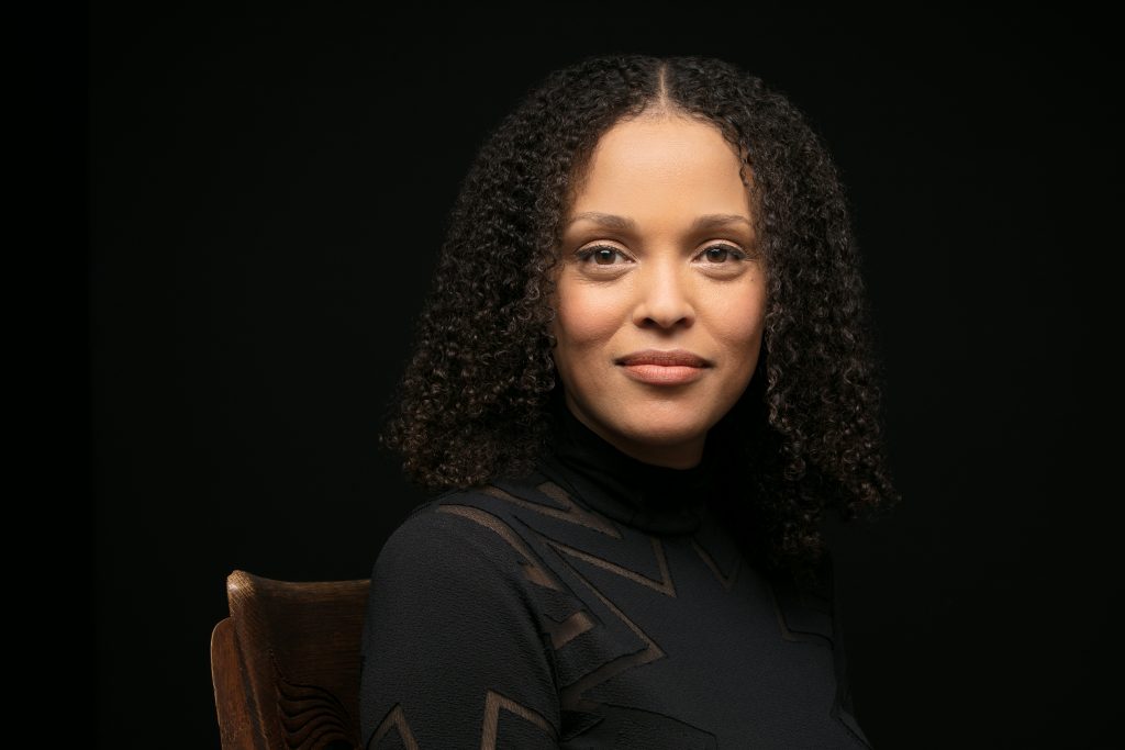 A bust photo of Jesmyn Ward. She is a light-skinned Black woman with tight shoulder-length ringlets and brown eyes. She is smiling peacefully at the camera and wearing a black blouse against a black background.