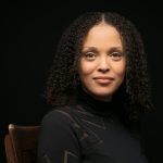 A bust photo of Jesmyn Ward. She is a lightskinned Black woman with tight shoulder-length ringlets and brown eyes. She is smiling peacefully at the camera and wearing a black blouse against a black background.