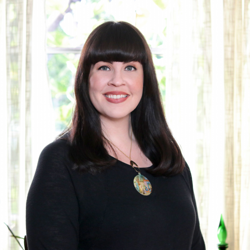 A bust portrait of Caitlin Doughty, a paleskinned woman with straight dark hair a bit past her shoulders and grey eyes. She is smiling at the camera.