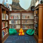 A photo of a reading nook lined with shelves and painted with a treehouse mural. Two green beanbag chairs and an inflatable campfire sit on the floor. At the top of the entryway is a swag of blue sheer fabric and light up stars.