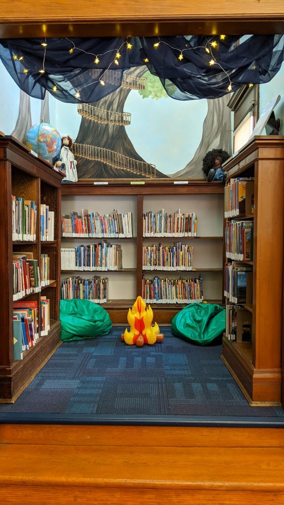 A photo of a reading nook lined with shelves and painted with a treehouse mural. Two green beanbag chairs and an inflatable campfire sit on the floor. At the top of the entryway is a swag of blue sheer fabric and light up stars.