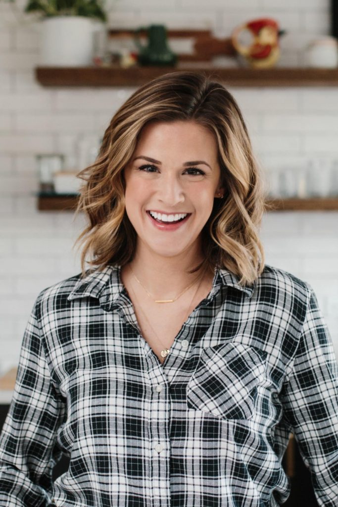 A portrait of Shauna Sever, who is a pale woman with dirty-blonde hair and brown eyes. She is wearing a grey plaid button down and is smiling at the camera.