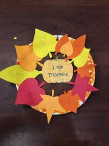 A "thankful wreath" made out of a paper plate and different colored paper. Paper leaves say different things the person is thankful for and in the middle is a pumpkin that says "I am thankful."